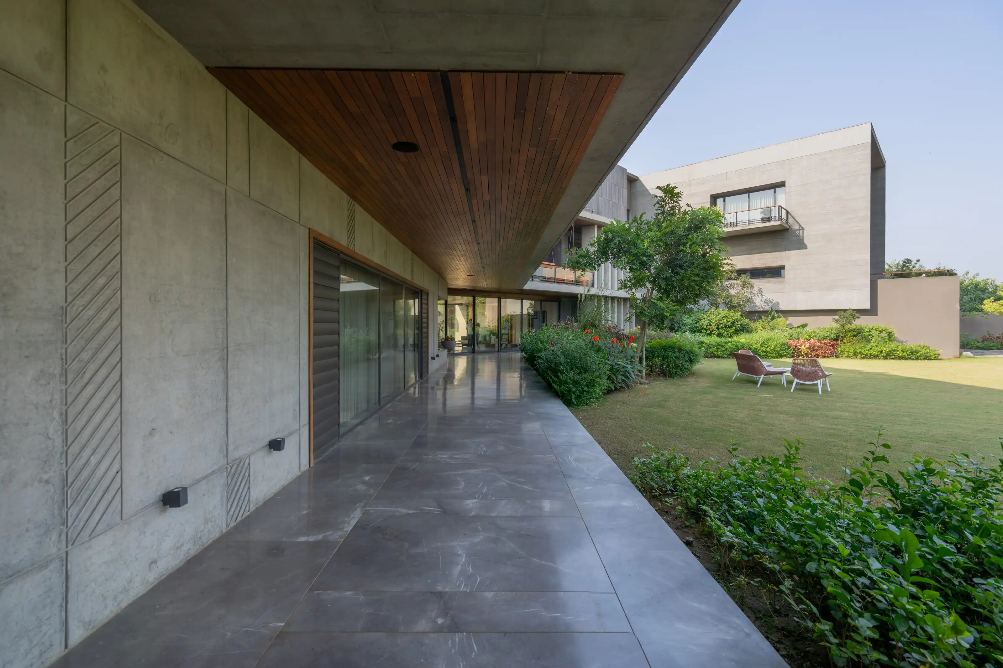 Board-formed concrete corridor overlooking a lawn with wicker chairs beneath a slatted wood soffit