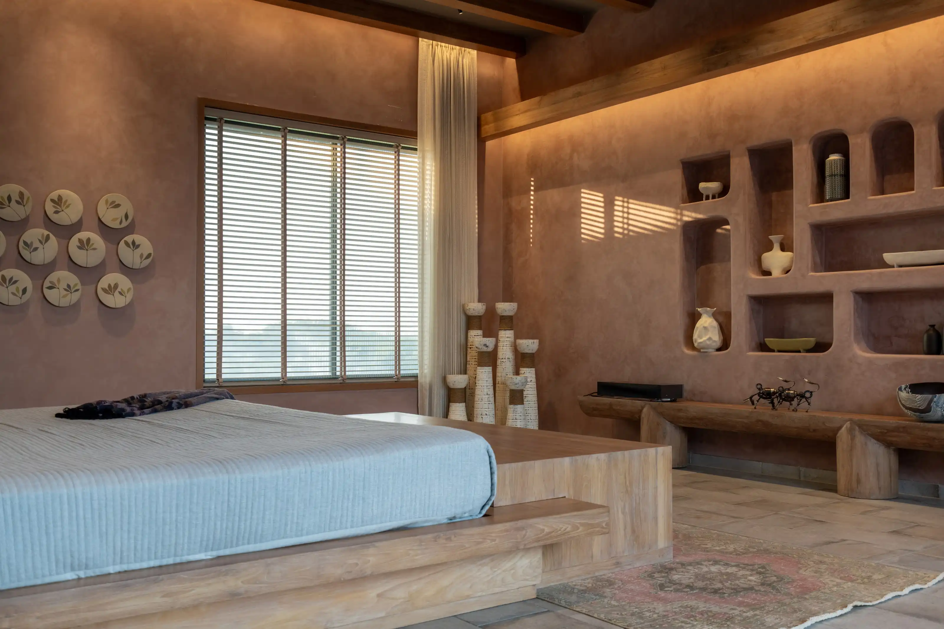 Terracotta-plastered bedroom with a floating timber bed and a wall of carved ceramic display niches