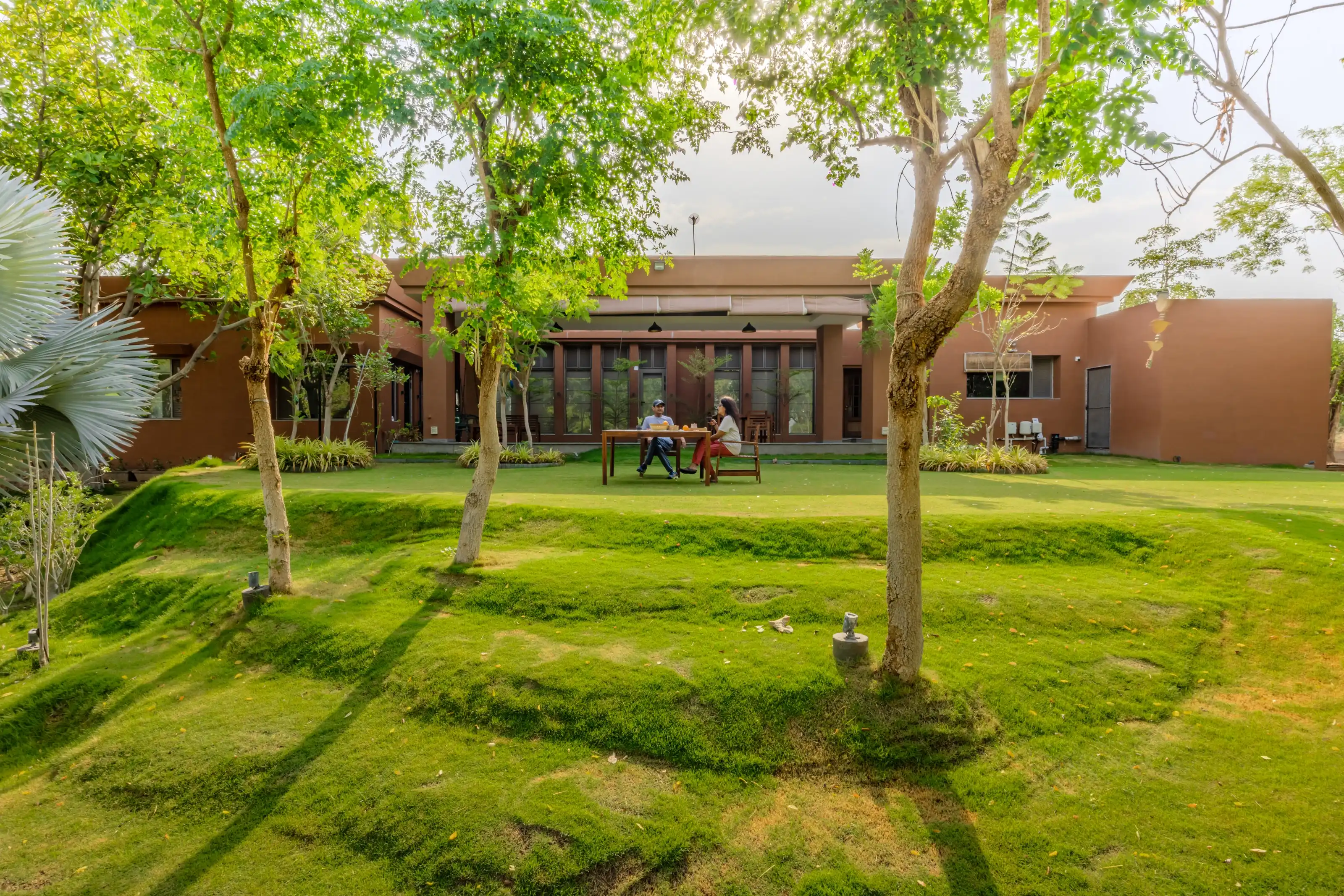Terracotta-plastered farmhouse facade with a couple seated at a garden table beneath mature trees