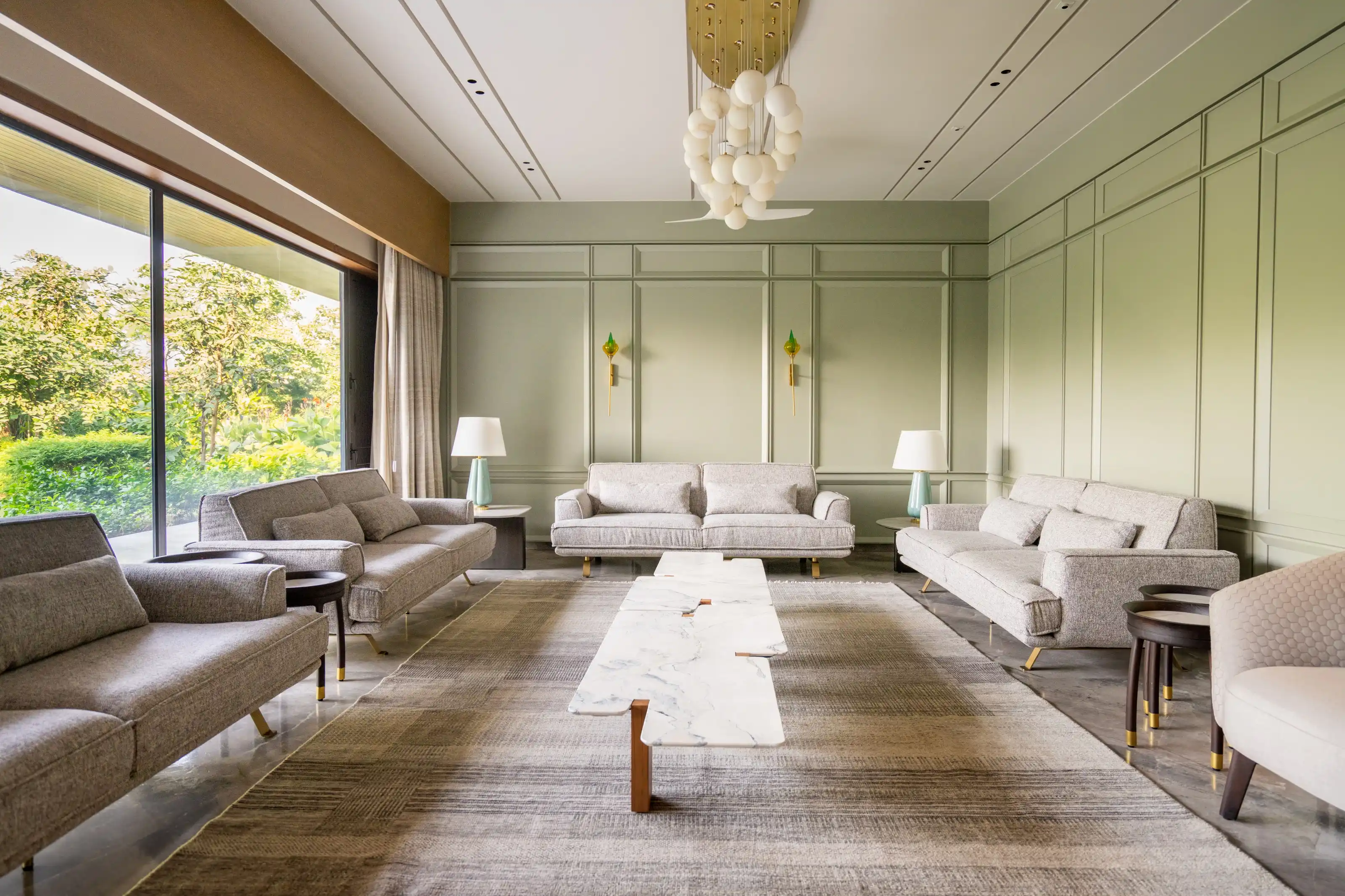Panelled sage-green living room with three grey sofas beneath a cluster chandelier of frosted globes