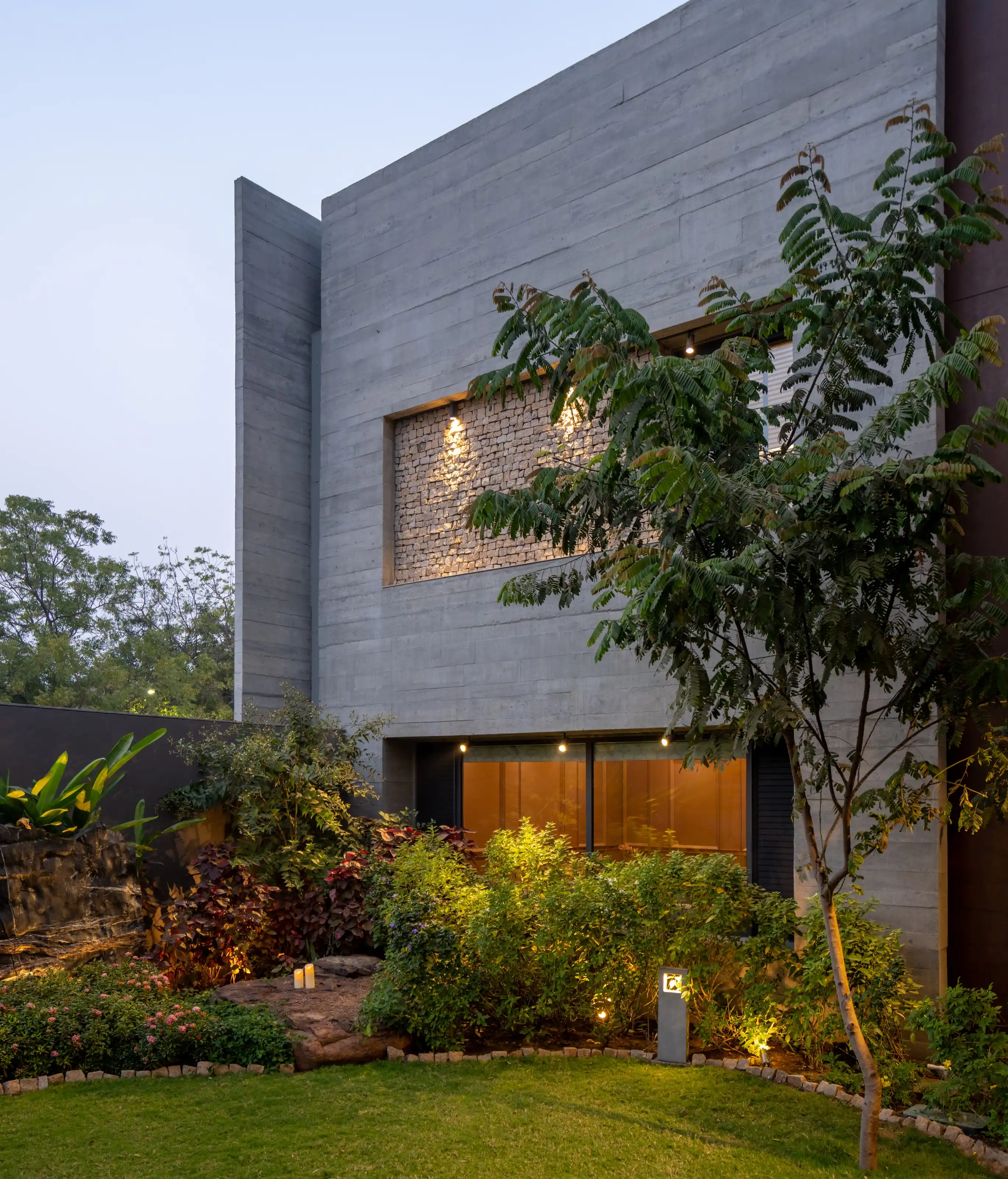 Dusk exterior of a board-formed concrete house with a raised stone accent panel and garden greenery
