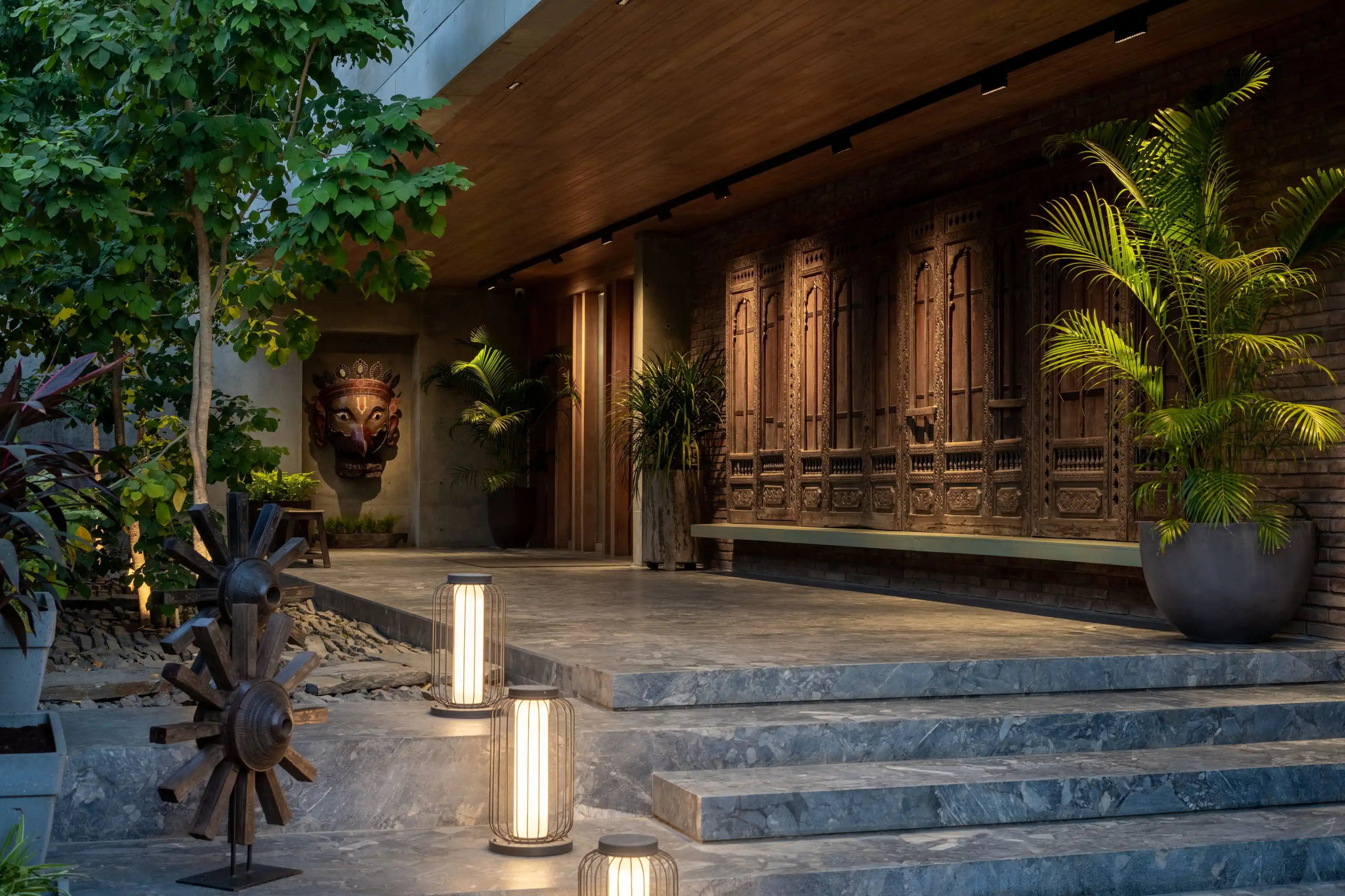 Covered entrance terrace at dusk with stone steps, lantern lights and carved timber panels set into a brick wall