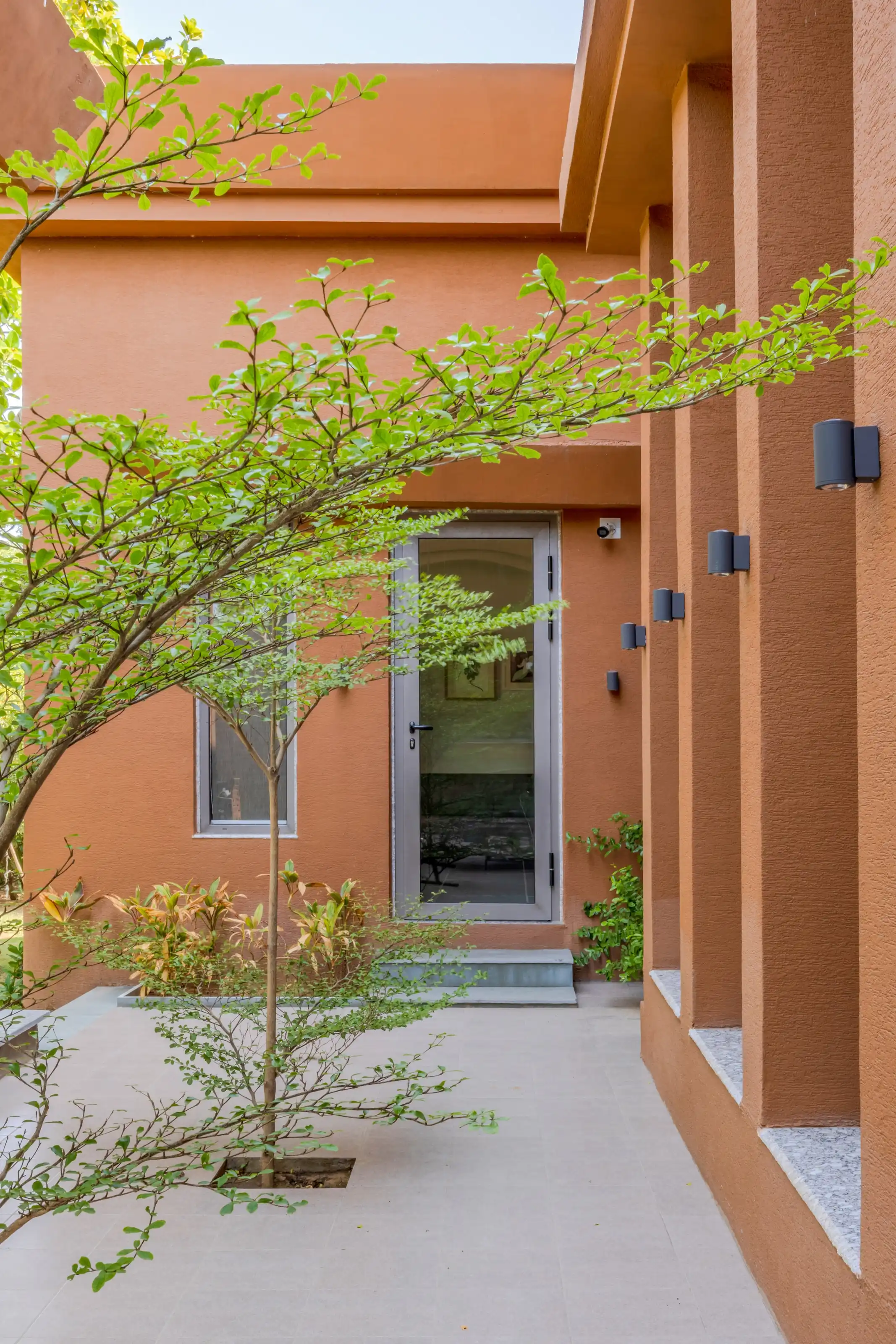 Covered walkway along the bedroom wing lit by a row of outdoor wall lamps beside a young tree